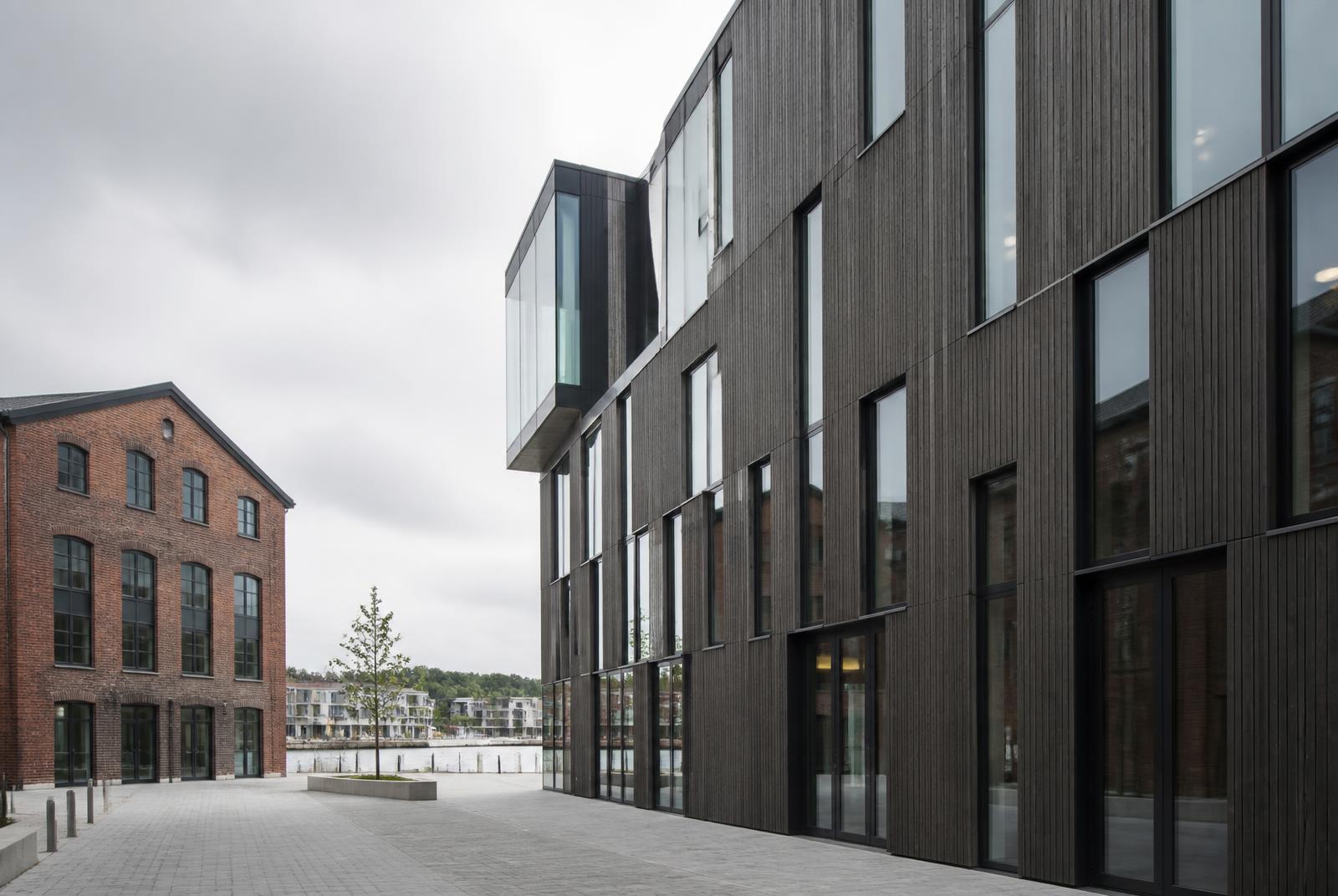 Nyere flerbrukerbygg i mørkt trepanel og glass ved siden av eldre teglbygg i et transformert sjønært næringsområde, relevant for kontorflytting til Verket i Moss